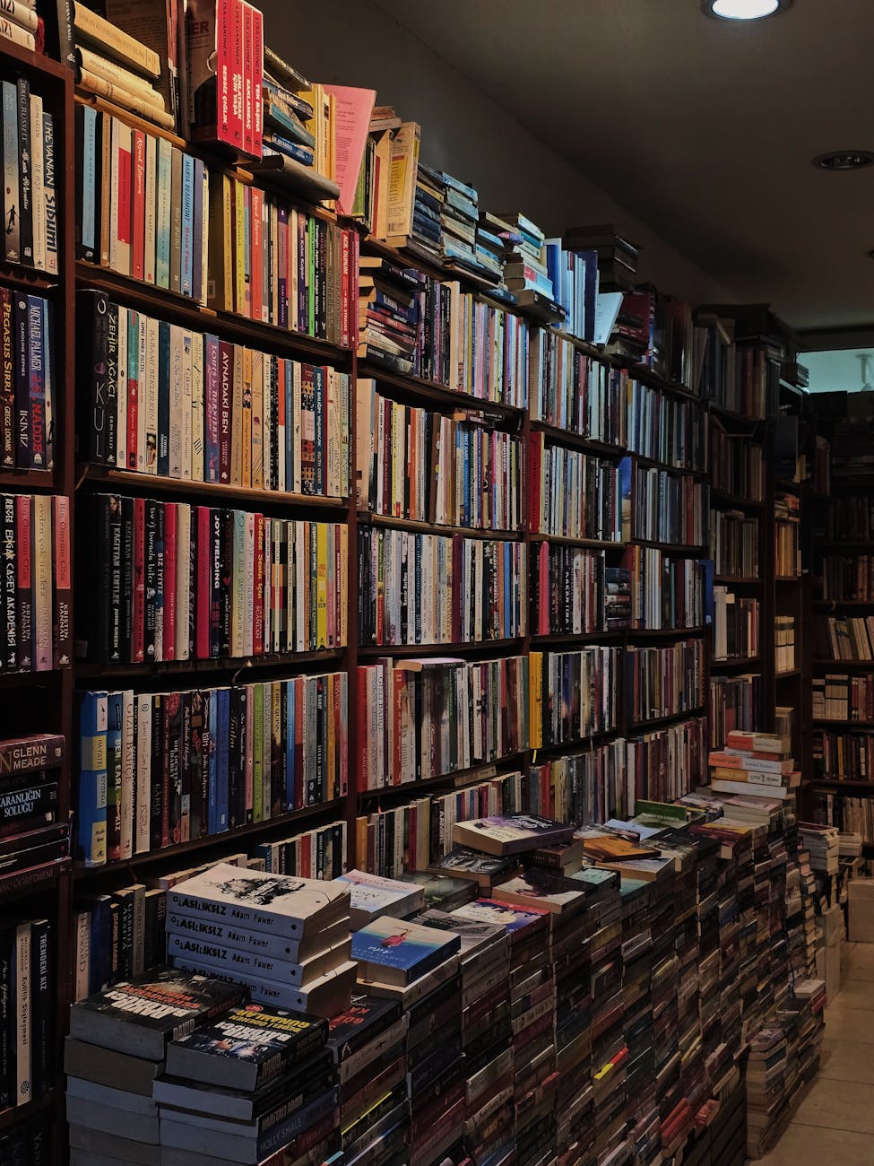 Tightly packed rows of books on shelves with some stacked on top of the shelf and many more stacked on the floor in front of the shelving.